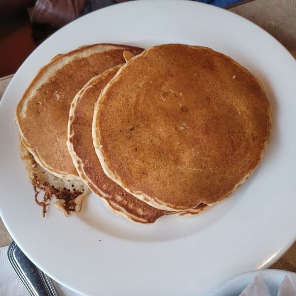 A stack of three golden-brown pancakes on a white plate, with a fork and napkin nearby.