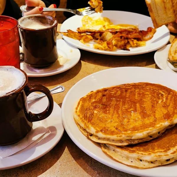 A table laden with breakfast dishes including pancakes, eggs, bacon, toast, and coffee.