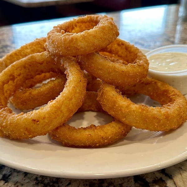 A plate of golden-brown onion rings with a side of creamy dipping sauce on a speckled countertop.